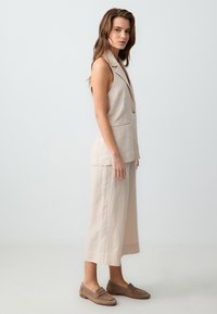 Lichtbeige linnen twee-delig kledingstuk met een mouwloze blazer met een zak en wijde pantalon. Bruine loafers maken de look compleet.