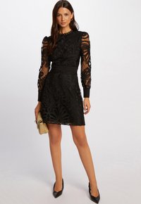 Robe en dentelle noire avec des manches longues, des motifs floraux intricats, une taille cintrée et une jupe évasée. Accentuée par une ceinture solide.