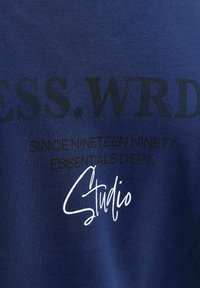 Navy blue fabric t-shirt featuring large black text reading "ESS. WRD." and smaller text "SINCE NINETEEN NINETY ESSENTIALS DEPT." with a white script "Studio."
