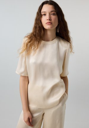 Jeune femme aux longs cheveux ondulés portant un haut court en satin crème et un pantalon assorti, debout, une main dans la poche.