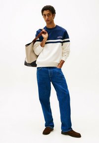 Felpa blu marino e crema con righe orizzontali, jeans in denim e mocassini marroni, mentre tiene una borsa tote beige con accenti scuri.