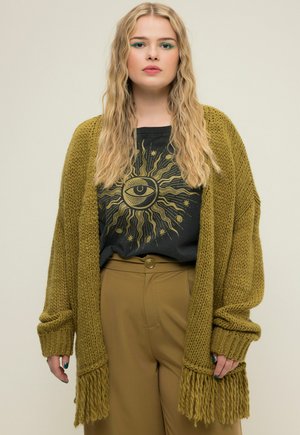 Olijfgroene oversized gebreide cardigan met franjes, gedragen over een zwart graphic t-shirt met een zon- en oogontwerp, gecombineerd met tan broek.