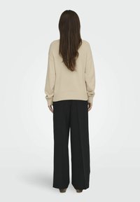 Beige, túlméretezett pulóver, bordás mandzsettákkal és aljjal, magas derékú, fekete, bő szárú nadrággal párosítva, hátulról nézve.