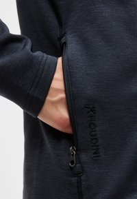 Χέρι μερικώς εισαγμένο στην τσέπη με φερμουάρ ενός σκούρου ναυτικού μακρυμάνικου ρούχου με κεντημένο το "HOUDINI" δίπλα στο φερμουάρ.