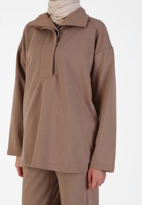 Losse, bruine pullover met een kleine kraag en een verborgen sluiting. Lange mouwen en een zachte textuur, gemaakt van rekbare stof.