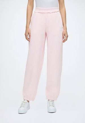 Personne portant un pantalon de survêtement ample rose clair et des baskets blanches, debout devant un fond uni.