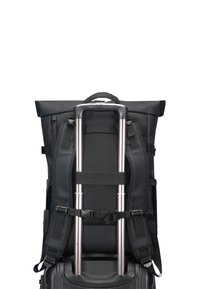 Schwarzer Rucksack, mit Gurten am ausgezogenen Griff eines schwarzen Rollkoffers befestigt, vor weißem Hintergrund.