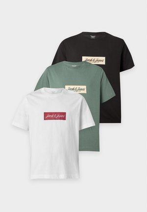 Mand iført en grøn t-shirt med logo, brune bukser og sorte sandaler mod en ensartet lys baggrund.