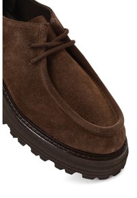 Brown suede lace-up shoe with thick textured black sole, rounded toe, and visible stitching along the edge.