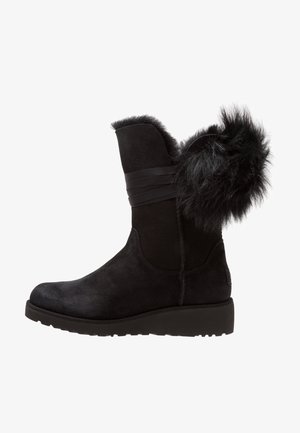 Botte d'hiver noire en daim mi-mollet avec garniture en fourrure et sangle décorative, semelle épaisse plate et détail en fourrure duveteuse à l'arrière.