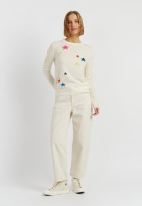 Maglione crema con motivi a forma di stella multicolori. Abbinato a pantaloni larghi beige chiaro e sneakers alte crema. Sfondo neutro.