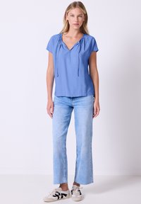 Donna in piedi contro uno sfondo bianco, indossa una blusa azzurra a maniche corte, jeans azzurri chiari e sneakers bianche con dettagli neri.