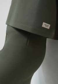 Abbigliamento verde, aderente, realizzato in tessuto morbido e testurizzato. L'etichetta riporta "ZERO WASTE DESIGN". Linee pulite ed elementi di design minimalista.