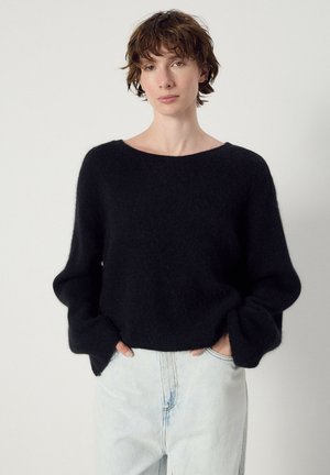 Young person with short brown hair wears a black fuzzy sweater and light blue jeans, standing against a plain light background.