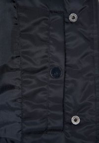 Svart vadderad tyg med en slät yta, som har två metallknappar och diskret sömdetalj längs kanterna.