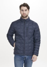 Marinblå vadderad jacka med dragkedja framtill, ståkrage och quiltad textur. Har sidofickor och en bröstficka för förvaring.