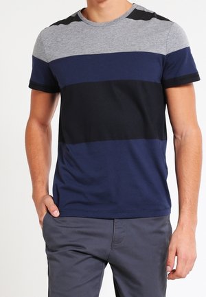 Mann trägt ein kurzärmeliges T-Shirt mit horizontalen grauen, marineblauen und schwarzen Streifen und graue Hose, steht mit einer Hand in der Tasche.