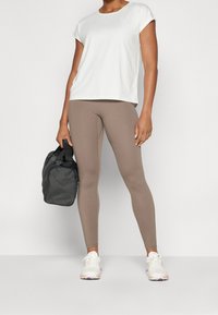 Camiseta deportiva blanca de manga corta combinada con leggings beige y zapatos blancos. Una bolsa negra se sostiene en una mano. Tela suave y diseño ajustado.