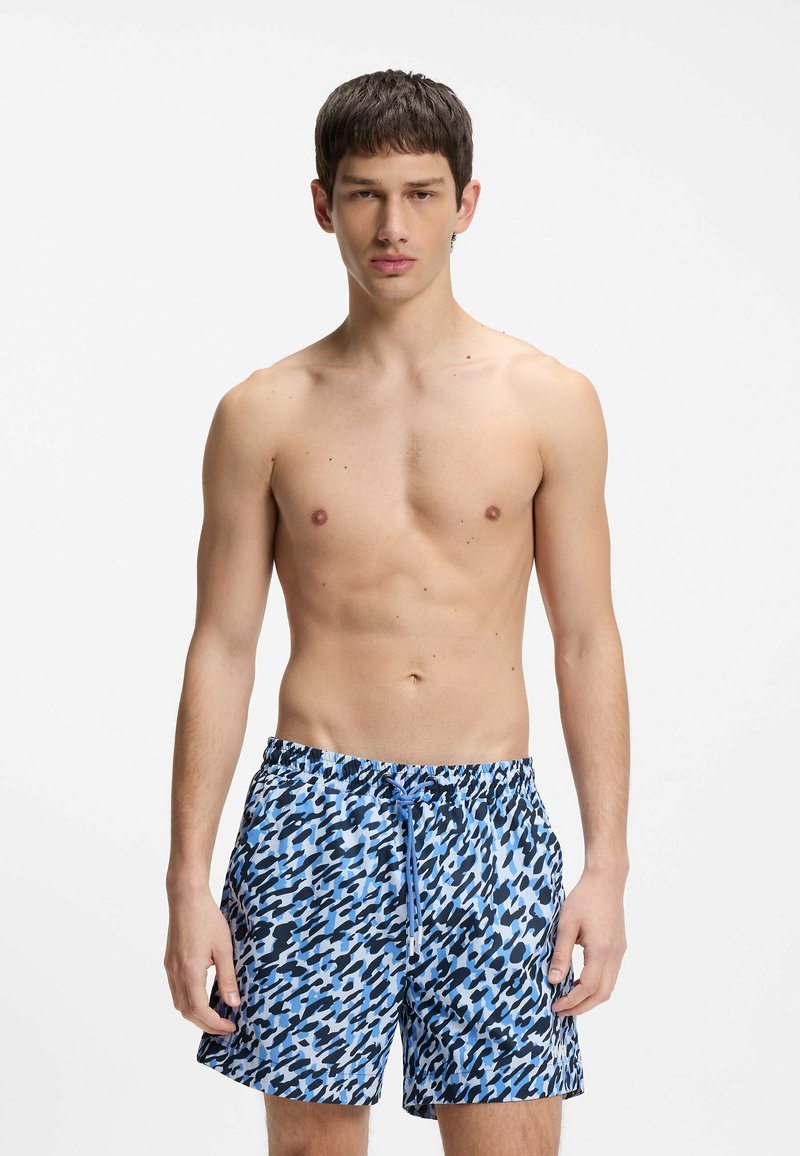 Jeune homme debout, torse nu, portant un short de bain bleu et noir à motifs avec un cordon, sur fond blanc.