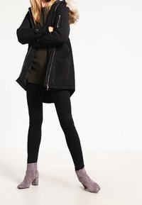 Černá parka bunda s umělým kožíškem na kapuci, nošená přes tmavě zelený svetr a černé legíny, doplněná šedými kotníkovými botami na podpatku.