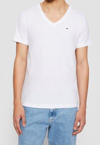 Bílé tričko s výstřihem do V vyrobené z bavlny, krátký rukáv, s malým logem na levém prsu, v kombinaci se světle modrými džíny.