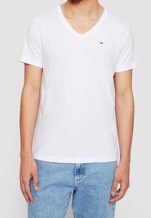 Camiseta blanca de cuello en V hecha de algodón, con mangas cortas, que presenta un pequeño logo en el lado izquierdo del pecho, combinada con vaqueros de color azul claro.
