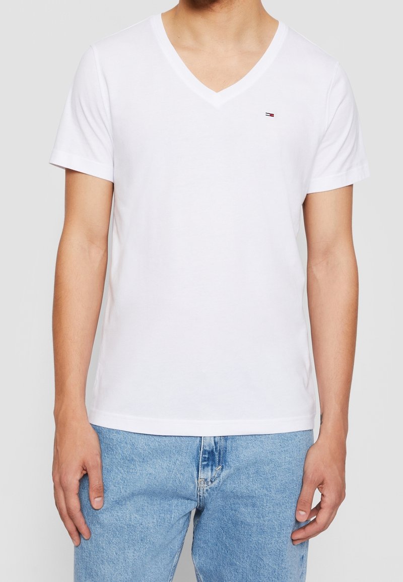 Bílé tričko s výstřihem do V vyrobené z bavlny, krátký rukáv, s malým logem na levém prsu, v kombinaci se světle modrými džíny.