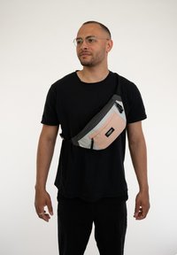 Borsa a tracolla frontale in grigio, rosa e nero. Presenta una tracolla regolabile, chiusura con zip e texture in tessuto. Indossata a tracollo.