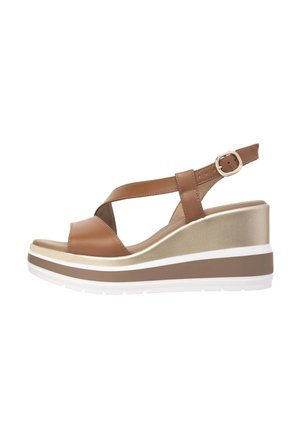 Brun syntetisk kile sandal med krydsede remme, guld accenter og hvid og brun lagdelt sål. Justerbar ankelrem for pasform.