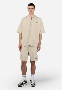 Beige kortærmet skjorte med et tekstureret mønster, knap-front design og diskret branding; parret med matchende shorts der har et logo.