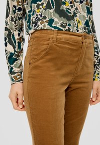 Bruine corduroy broek met een soepele textuur, voorzien van zijzakken en contrasterende stiksels. Draagt een gestreept shirt met lange mouwen.