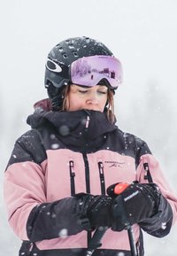 Kurtka narciarska w różowo-czarnym wzorze blokowym, wykonana z ocieplanej tkaniny. Akcesoria obejmują czarny kask i odblaskowe gogle.