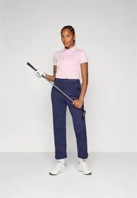 Golfkläder består av en ljusrosa pikétröja och marinblå byxor. Personen håller i en golfklubba, bär vita sneakers och en golfhandske.