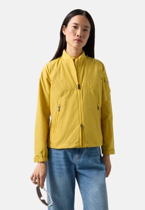 Frau mit langen dunklen Haaren, die eine gelbe Reißverschlussjacke und blaue Jeans trägt und eine Sonnenbrille in der rechten Hand vor weißem Hintergrund hält.