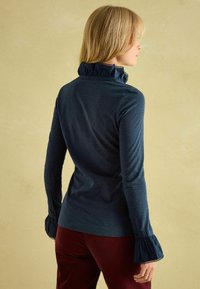 Navyblaues Langarmoberteil mit Rüchenkragen, feinen vertikalen Streifen und ausladenden Manschetten. Der Stoff wirkt glatt mit einem leichten Glanz.