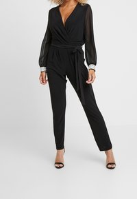 Svart jumpsuit med omslagdesign, genomskinliga långärmar och silverdetaljer på ärmsluten. Har knytskärp i midjan och kroppsnära ben.