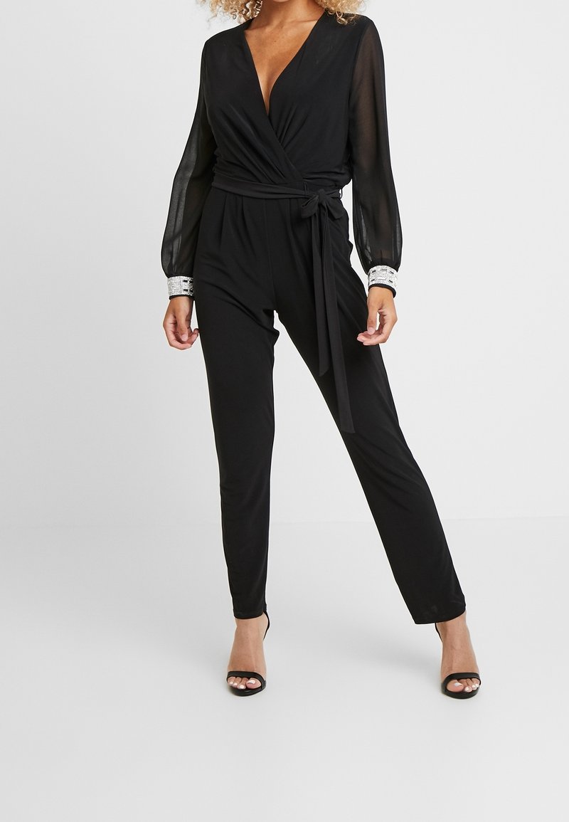 Svart jumpsuit med omslagdesign, genomskinliga långärmar och silverdetaljer på ärmsluten. Har knytskärp i midjan och kroppsnära ben.