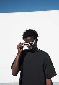 Črna površna majica, mat bele angularne sončne očala s temnimi lečami, teksturirane lase, minimalistična ozadje. Svetla in jasna osvetlitev.