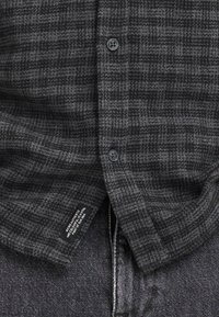 Tamnosiva i crna karirana košulja s gumbima, djelomično prekriva tamne traperice s vidljivim područjem munje i gumba.