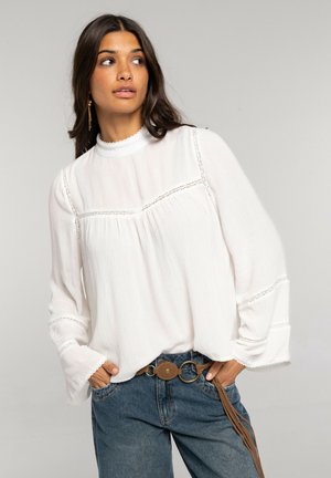 Mujer con cabello largo y oscuro que lleva una blusa blanca con detalles de encaje y mangas acampanadas, vaqueros azules y un cinturón marrón con hebillas circulares.