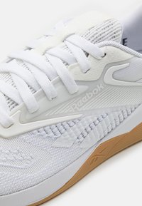 Λευκά αθλητικά παπούτσια Reebok με υφασμάτινη επένδυση, λευκές κορδόνες και σόλα από καουτσούκ γκόμε. Το λογότυπο της μάρκας φαίνεται στο πλάι.