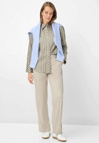 Camicia a righe verde oliva, pantaloni beige, maglione azzurro chiaro drappeggiato sulle spalle, scarpe da ginnastica bianche, design minimalista e colori neutri.