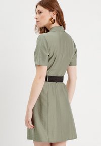 Kurzärmeliges Kleid in hellolivgrün, mit einer taillierten Taille und einem schwarzen, strukturierten Gürtel. Glattes Material mit einem Hauch von Glanz.
