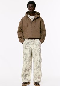 Mand står med hænderne i lommerne iført en brun zip-up hættetrøje, hvide og brune mønstrede cargo bukser, perlekæde og brune sko.