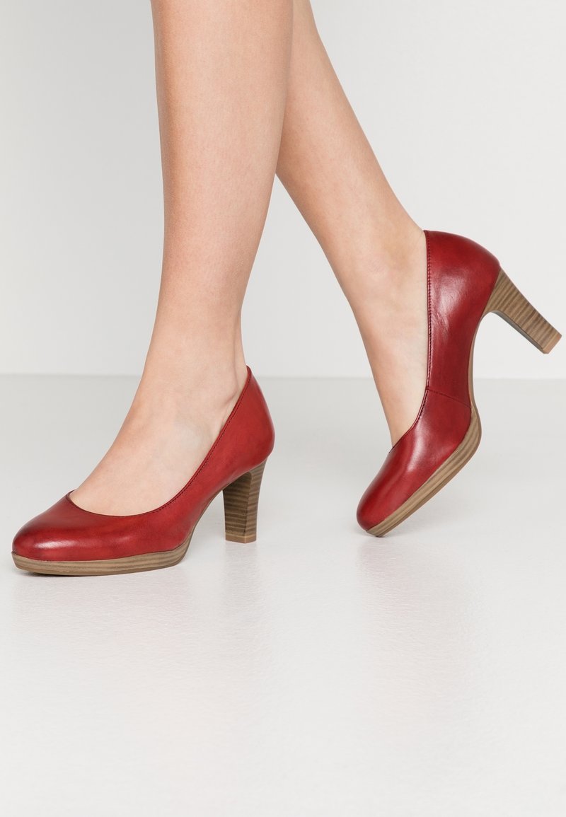 Jambes portant des escarpins rouges en cuir à talons moyens avec talons blocs en bois, debout sur un sol lisse blanc contre un fond uni.