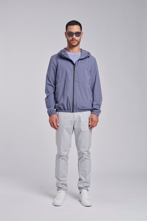 Mężczyzna w niebieskiej kurtce z kapturem, jasnoszarych spodniach, białych trampkach i ciemnych okularach przeciwsłonecznych stoi z rękami swobodnie opartymi na prostym, szarym tle.