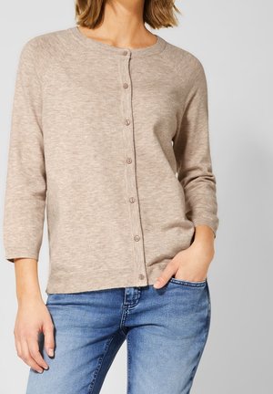 Vrouw die een beige, lange mouwen, knoopjesdichte cardigan met een ronde hals en blauwe spijkerbroek draagt tegen een effen achtergrond.