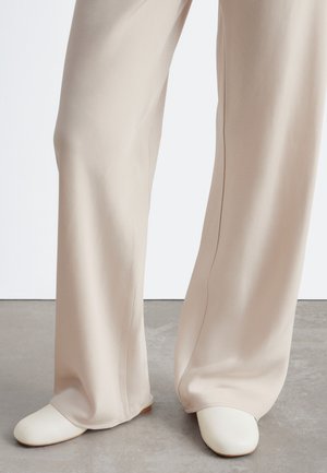 Pantalon beige à jambes larges porté avec des chaussures plates blanches à bout arrondi sur un sol gris sur fond blanc.