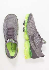 Szare i żółte buty Nike Vapormax z dzianinową cholewką, zielonymi akcentami, przezroczystymi podeszwami powietrznymi i czarną gumową podeszwą. Lekka konstrukcja.
