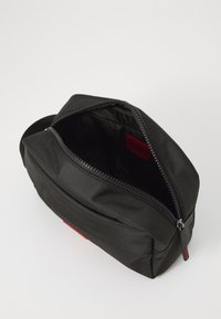 Svart nylon toalettmappe med glidelås, med en rød innvendig etikett og en teksturert utvendig overflate.